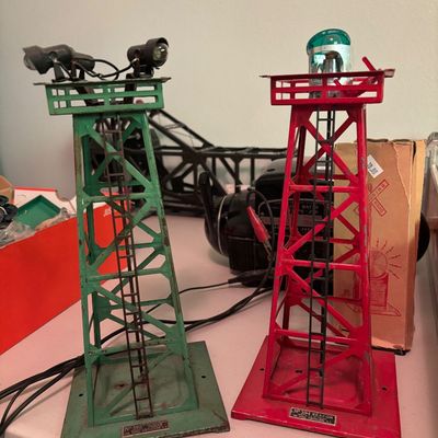 Vintage Lionel O gauge train accessories, featuring a green No. 395 floodlight tower and a red No. 394 rotary beacon tower, both showing...