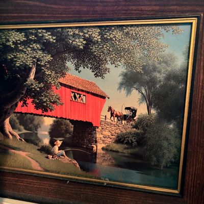 Framed vintage print depicting a nostalgic rural scene with a red covered bridge, a horse-drawn carriage, and a boy fishing by the river.