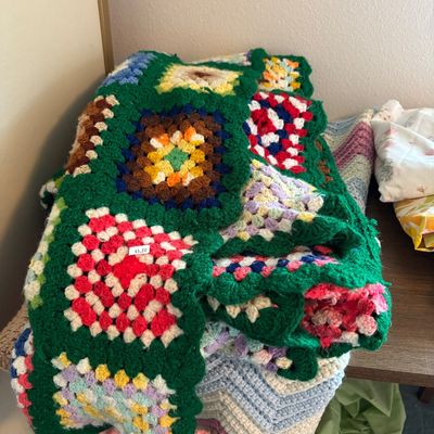 Vintage handmade crochet granny square blanket with a vibrant green border and multi-colored centers, stacked on top of other knit blankets.