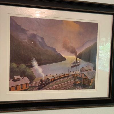 Framed and matted art print depicting a vintage steam locomotive and a steamboat at a mountain lake depot, housed in a dark wooden frame.