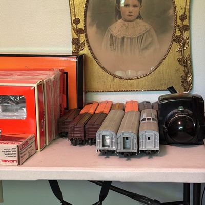Vintage Lionel model train cars, track accessories, and original boxes, displayed alongside an antique oval-framed portrait of a young girl.