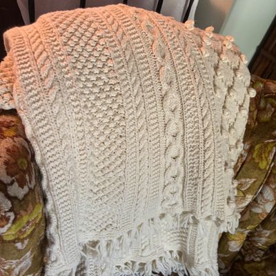 Vintage cream-colored knit throw blanket featuring intricate cable knit patterns and fringed edges, draped over an armchair.
