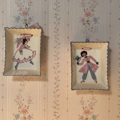Pair of vintage ceramic wall plaques with scalloped edges, featuring hand-painted illustrations of a Victorian-style lady and gentleman...