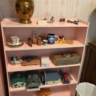 Vintage pink painted wooden bookshelf holding various collectibles, including a brass table lamp, small figurines, teacups, and toy houses.