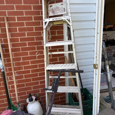 Vintage aluminum step ladder showing signs of wear and use, alongside various yard tools and a garden sprayer.