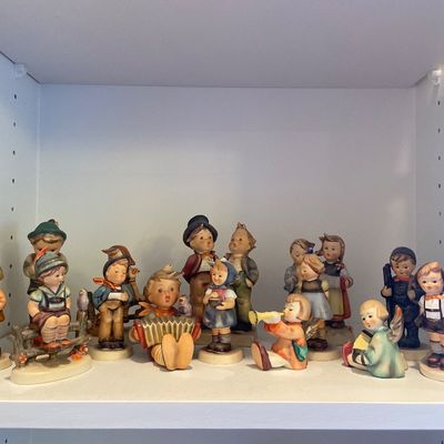 Sale Photo Thumbnail #130: Collection of vintage Goebel Hummel porcelain figurines featuring children playing musical instruments, singing, and posing on a white display shelf.