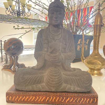 Sale Photo Thumbnail #48: Carved stone seated Buddha statue displayed on a patterned wooden plinth, flanked by decorative brass objects and a polished stone sphere.