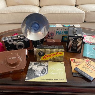Sale Photo Thumbnail #121: Vintage photography equipment collection featuring an Argus C-3 35mm rangefinder camera with leather case and manual, a Kodak Brownie Target Six-20 box camera, flash attachment, bulbs, and related ephemera.