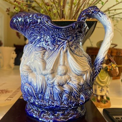 Sale Photo Thumbnail #105: Vintage cobalt blue and white embossed potteryp pitcher featuring detailed figurative scenes in high relief, displayed on a wooden stand.