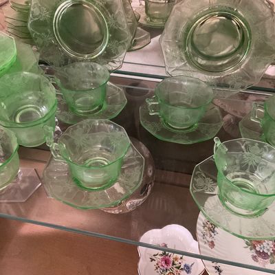 Sale Photo Thumbnail #8: Collection of green depression glass teacups, saucers, and plates featuring an etched floral pattern, displayed on a glass shelf.