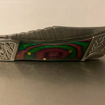 Sale Photo Thumbnail #75: Folding pocket knife featuring a Damascus steel blade, engraved metal bolsters, and a multi-colored laminated wood handle scale in green, purple, and red.