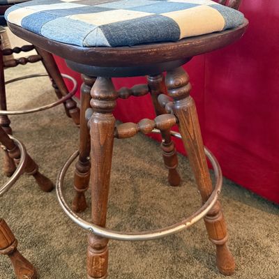 Sale Photo Thumbnail #249: A set of four vintage wooden swivel bar stools featuring a classic spindle back design and a circular metal footrest. The seats have been upholstered in a unique blue and grey denim patchwork fabric. The stools show some light wear on the wooden legs and 