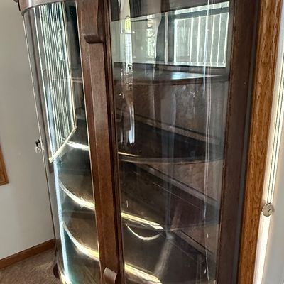 Sale Photo Thumbnail #185: An antique tiger oak china display cabinet featuring a curved glass front door and curved glass side panels. The cabinet includes five wooden shelves, a mirrored upper back section, and elegant carved wood details. It is fitted with a vintage brass and fa