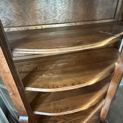 Sale Photo Thumbnail #184: An antique tiger oak china display cabinet featuring a curved glass front door and curved glass side panels. The cabinet includes five wooden shelves, a mirrored upper back section, and elegant carved wood details. It is fitted with a vintage brass and fa