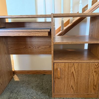 Sale Photo Thumbnail #231: A vintage-style laminate wood computer desk featuring an oak grain finish. Designed with a slide-out keyboard tray on metal tracks, open shelving compartments, and a lower storage cabinet with a single door. Ideal for small spaces, home offices, or studen