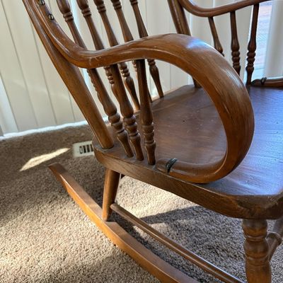 Sale Photo Thumbnail #207: A vintage wooden rocking chair featuring a detailed carved 'North Wind' or green man face on the top crest rail. The chair is constructed with turned spindle supports, curved armrests, and a contoured seat. It shows signs of wear consistent with age and u