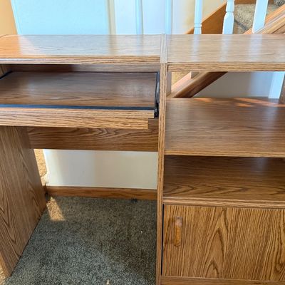 Sale Photo Thumbnail #232: A vintage-style laminate wood computer desk featuring an oak grain finish. Designed with a slide-out keyboard tray on metal tracks, open shelving compartments, and a lower storage cabinet with a single door. Ideal for small spaces, home offices, or studen