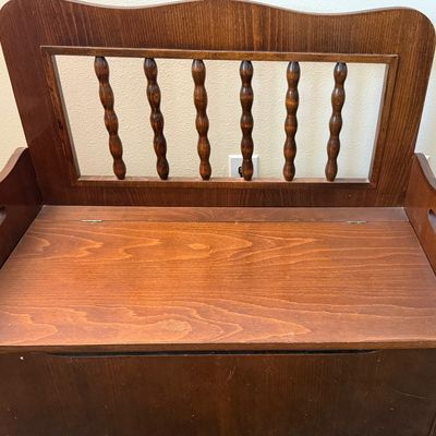 Sale Photo Thumbnail #237: A vintage wooden storage bench featuring a decorative spindle backrest, side handle cutouts, and a hinged seat that opens to a spacious storage compartment. The piece shows some light surface wear, minor scratches, and scuffs consistent with age and use. 