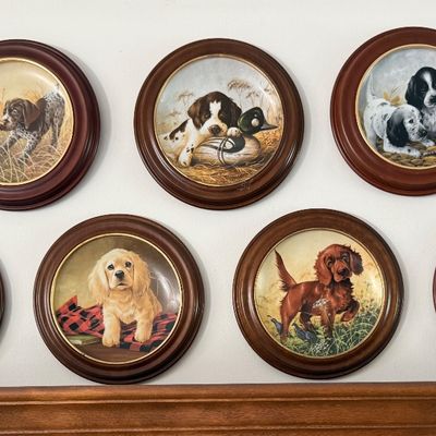 Sale Photo Thumbnail #46: A charming collection of seven vintage porcelain collector plates featuring various dog breeds and puppies in outdoor sporting scenes. Each plate is mounted in a round, dark wood frame with a gold-toned inner bezel. The artwork depicts breeds such as Gold