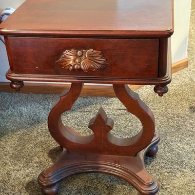 Sale Photo Thumbnail #84: A vintage wooden side table featuring a decorative lyre-shaped base, a single drawer with a carved leaf pull, and turned drop finials. The table top shows some light surface wear and minor scuffs consistent with age and use. Measures approximately 16.5 in