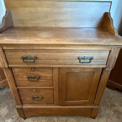 Sale Photo Thumbnail #168: An antique wooden washstand cabinet featuring a top gallery splashback, one wide top drawer, two smaller side drawers with keyholes, and a single cabinet door. The drawers showcase classic dovetail construction and are fitted with ornate metal pull handle