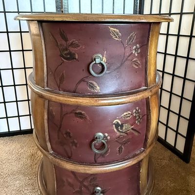 Sale Photo Thumbnail #175: A charming round wooden accent chest or nightstand featuring three curved drawers. The drawer fronts are finished in a deep burgundy/mauve tone and hand-painted with delicate bird and floral branch motifs. Features metal ring pull hardware. Shows some lig