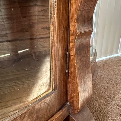 Sale Photo Thumbnail #192: An antique tiger oak china display cabinet featuring a curved glass front door and curved glass side panels. The cabinet includes five wooden shelves, a mirrored upper back section, and elegant carved wood details. It is fitted with a vintage brass and fa