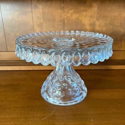 Sale Photo Thumbnail #48: A vintage clear pressed glass pedestal cake stand featuring the classic Fostoria 'American' 3D cube pattern. Features a scalloped, draped rim and a textured, flared pedestal base. Excellent condition with no visible chips, cracks, or clouding. Measures ap