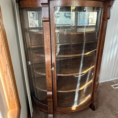 Sale Photo Thumbnail #181: An antique tiger oak china display cabinet featuring a curved glass front door and curved glass side panels. The cabinet includes five wooden shelves, a mirrored upper back section, and elegant carved wood details. It is fitted with a vintage brass and fa
