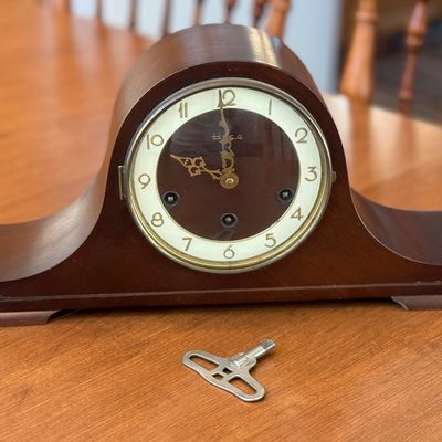 Sale Photo Thumbnail #15: A vintage Heco mechanical mantel clock featuring a classic wooden tambour-style case. The clock face has a two-tone design with a dark wood center, cream-colored outer ring, gold-tone Arabic numerals, and ornate hands. Includes three winding arbors on the