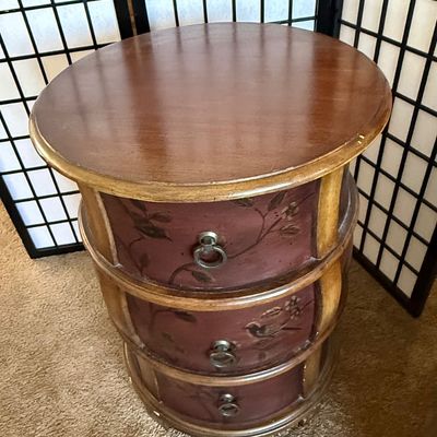 Sale Photo Thumbnail #176: A charming round wooden accent chest or nightstand featuring three curved drawers. The drawer fronts are finished in a deep burgundy/mauve tone and hand-painted with delicate bird and floral branch motifs. Features metal ring pull hardware. Shows some lig