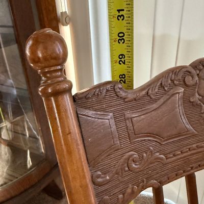 Sale Photo Thumbnail #208: A vintage wooden rocking chair featuring a detailed carved 'North Wind' or green man face on the top crest rail. The chair is constructed with turned spindle supports, curved armrests, and a contoured seat. It shows signs of wear consistent with age and u