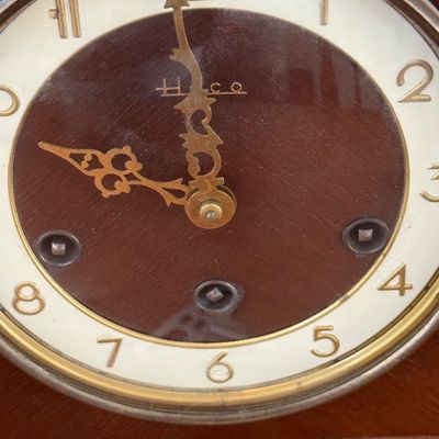 Sale Photo Thumbnail #16: A vintage Heco mechanical mantel clock featuring a classic wooden tambour-style case. The clock face has a two-tone design with a dark wood center, cream-colored outer ring, gold-tone Arabic numerals, and ornate hands. Includes three winding arbors on the