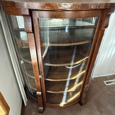 Sale Photo Thumbnail #182: An antique tiger oak china display cabinet featuring a curved glass front door and curved glass side panels. The cabinet includes five wooden shelves, a mirrored upper back section, and elegant carved wood details. It is fitted with a vintage brass and fa