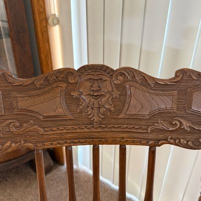 Sale Photo Thumbnail #206: A vintage wooden rocking chair featuring a detailed carved 'North Wind' or green man face on the top crest rail. The chair is constructed with turned spindle supports, curved armrests, and a contoured seat. It shows signs of wear consistent with age and u