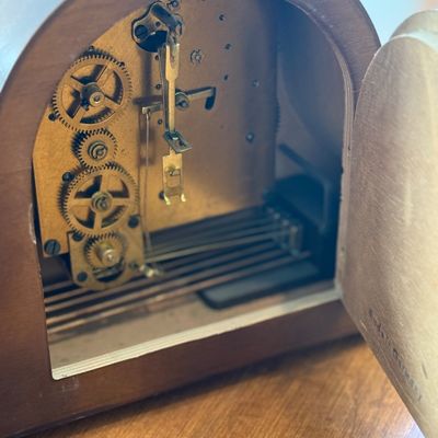 Sale Photo Thumbnail #17: A vintage Heco mechanical mantel clock featuring a classic wooden tambour-style case. The clock face has a two-tone design with a dark wood center, cream-colored outer ring, gold-tone Arabic numerals, and ornate hands. Includes three winding arbors on the