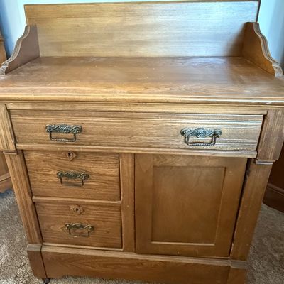 Sale Photo Thumbnail #169: An antique wooden washstand cabinet featuring a top gallery splashback, one wide top drawer, two smaller side drawers with keyholes, and a single cabinet door. The drawers showcase classic dovetail construction and are fitted with ornate metal pull handle