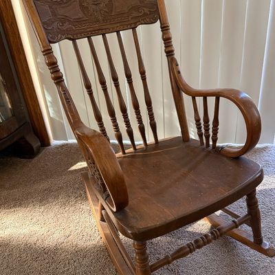 Sale Photo Thumbnail #205: A vintage wooden rocking chair featuring a detailed carved 'North Wind' or green man face on the top crest rail. The chair is constructed with turned spindle supports, curved armrests, and a contoured seat. It shows signs of wear consistent with age and u