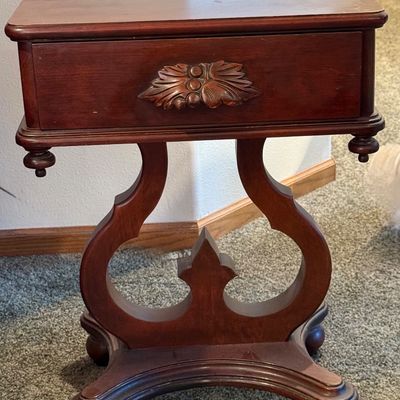 Sale Photo Thumbnail #83: A vintage wooden side table featuring a decorative lyre-shaped base, a single drawer with a carved leaf pull, and turned drop finials. The table top shows some light surface wear and minor scuffs consistent with age and use. Measures approximately 16.5 in