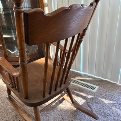 Sale Photo Thumbnail #210: A vintage wooden rocking chair featuring a detailed carved 'North Wind' or green man face on the top crest rail. The chair is constructed with turned spindle supports, curved armrests, and a contoured seat. It shows signs of wear consistent with age and u