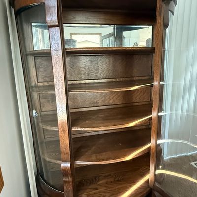 Sale Photo Thumbnail #183: An antique tiger oak china display cabinet featuring a curved glass front door and curved glass side panels. The cabinet includes five wooden shelves, a mirrored upper back section, and elegant carved wood details. It is fitted with a vintage brass and fa