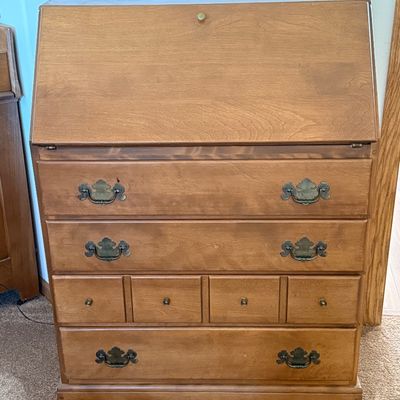 Sale Photo Thumbnail #161: A vintage wooden slant-front secretary desk featuring a drop-down writing surface that opens to reveal an interior organizer with small drawers and mail slots. The lower section includes three large drawers and a row of four smaller drawers, all accented 