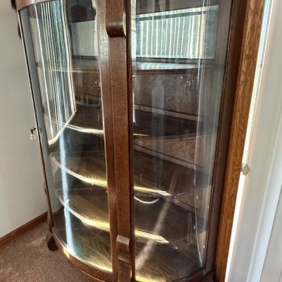 Sale Photo Thumbnail #186: An antique tiger oak china display cabinet featuring a curved glass front door and curved glass side panels. The cabinet includes five wooden shelves, a mirrored upper back section, and elegant carved wood details. It is fitted with a vintage brass and fa