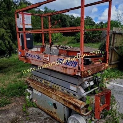 Sale Photo Thumbnail #41: Skyjack SJIII 3219 electric scissor lift showing significant weathering, rust, and overgrowth from outdoor storage.
