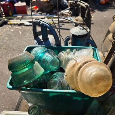 Sale Photo Thumbnail #23: Collection of vintage glass electrical insulators in various shades of green and clear glass, displayed in a green plastic bin alongside old metal lanterns.