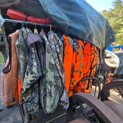 Sale Photo Thumbnail #2: Assortment of outdoor hunting apparel, including camouflage jackets and high-visibility blaze orange vests, hanging on a rack outdoors.