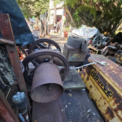 Sale Photo Thumbnail #3: Vintage hit-and-miss engine with large cast-iron flywheels and a flat belt pulley, showing significant surface rust and wear.