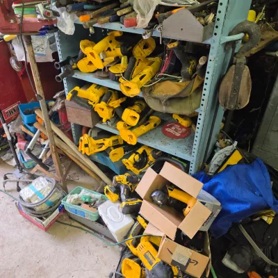 Sale Photo Thumbnail #61: Assortment of used DeWalt power tools, including reciprocating saws, drills, and circular saws, stored on a blue metal shelving unit in a workshop.