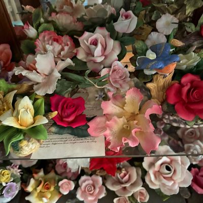 Sale Photo Thumbnail #55: Collection of vintage porcelain floral figurines, featuring detailed roses, orchids, and a bluebird, displayed on glass shelves.