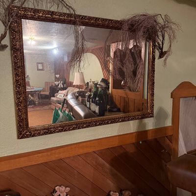 Sale Photo Thumbnail #33: Large rectangular wall mirror featuring an ornate, heavily detailed gold-finished frame, shown mounted above wood paneling.